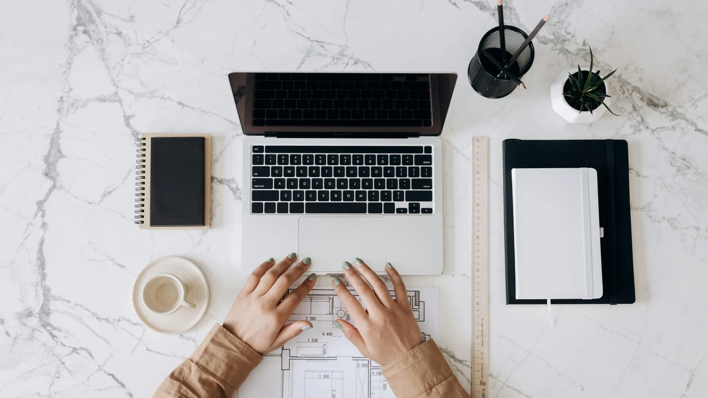 Overhead view of hands typing on a laptop at a marble desk beside a notebook and floor plans